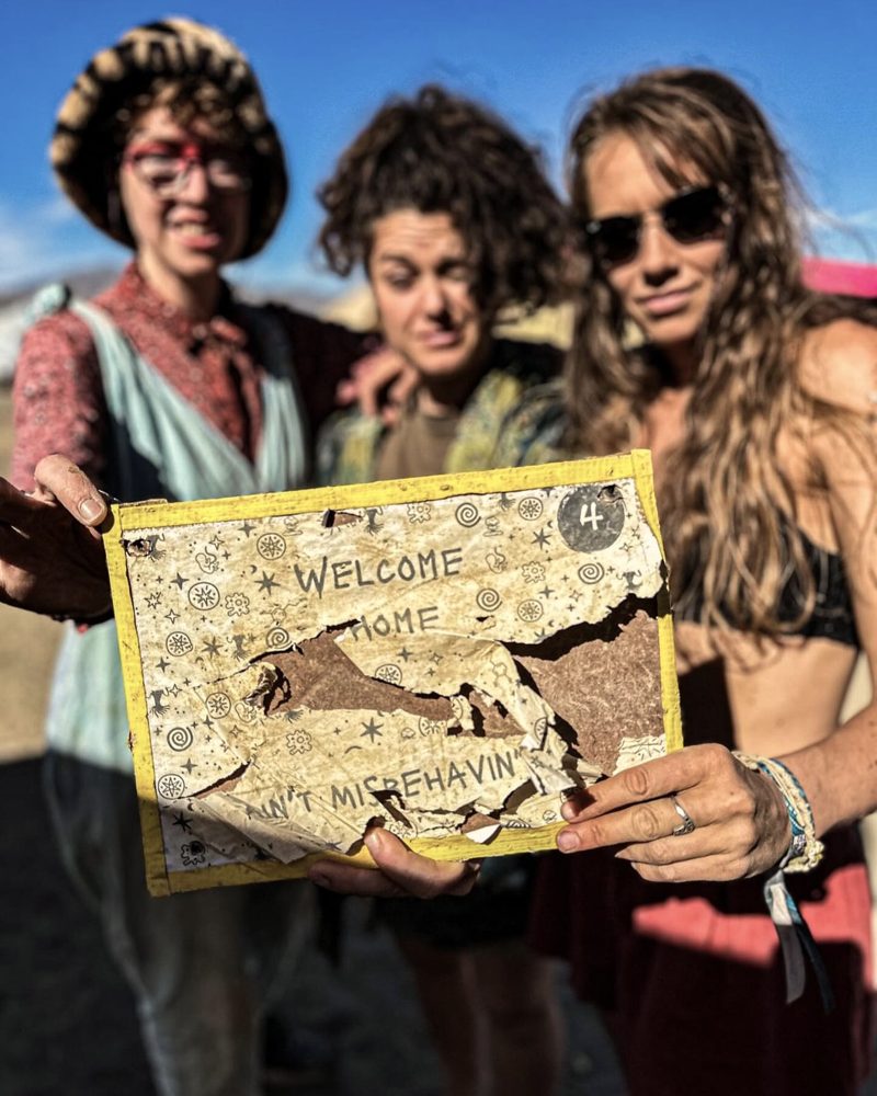 Muriel and two friends at AfrikaBurn holding a battered Ain't Misbehavin' welcome sign